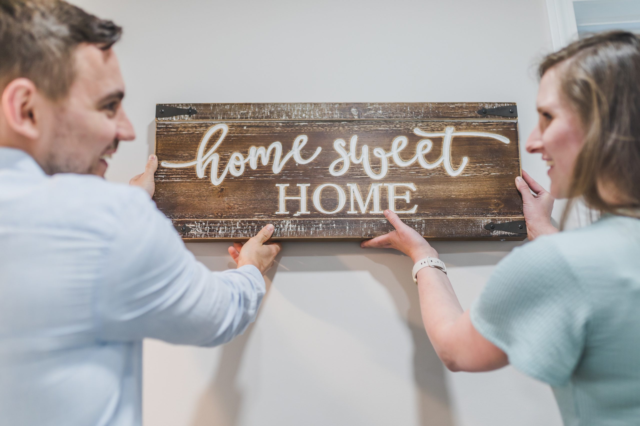 A man and a woman are hanging a wooden sign on a wall that reads “Home Sweet Home.” They are smiling and appear to be in a happy, domestic setting.